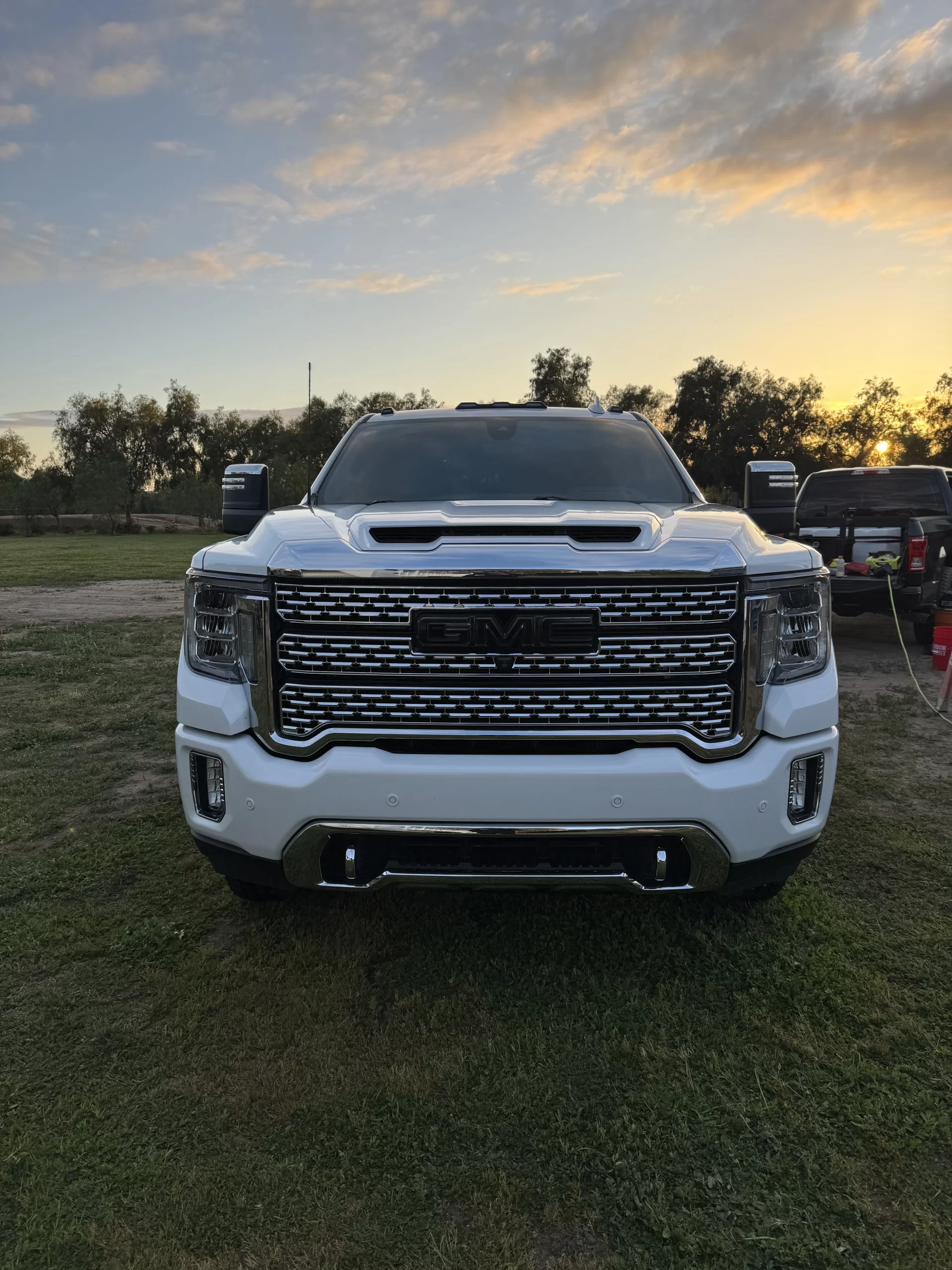 A white GMC pickup truck is parked on grass at sunset, viewed from the front. Trees, a fence, and another vehicle are visible in the background under a partly cloudy sky. - DNC Auto Detailing in Ramona, California