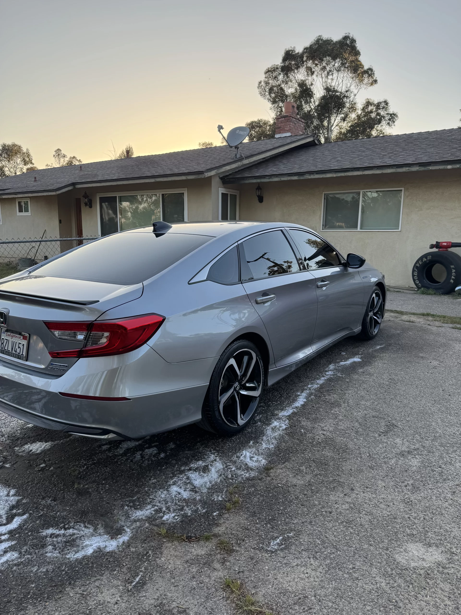 A silver sedan is parked in the driveway of a single-story house at sunset. The car has tinted windows and black alloy wheels. Trees and a tire are visible in the background. - DNC Auto Detailing in Ramona, California