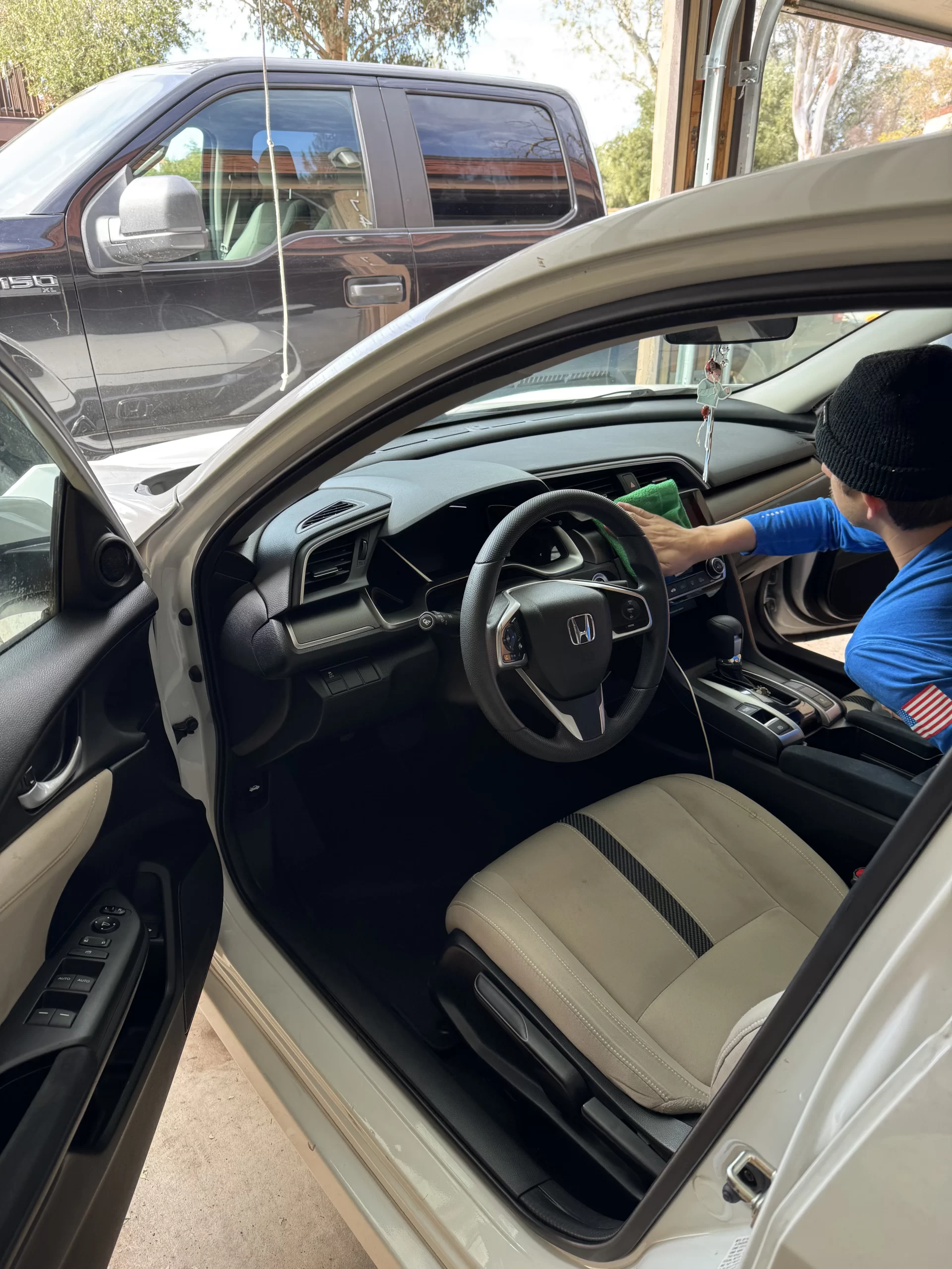 A person cleans the dashboard of a white Honda car with a green cloth. The driver's door is open, and a large black pickup truck is parked outside in the background. - DNC Auto Detailing in Ramona, California