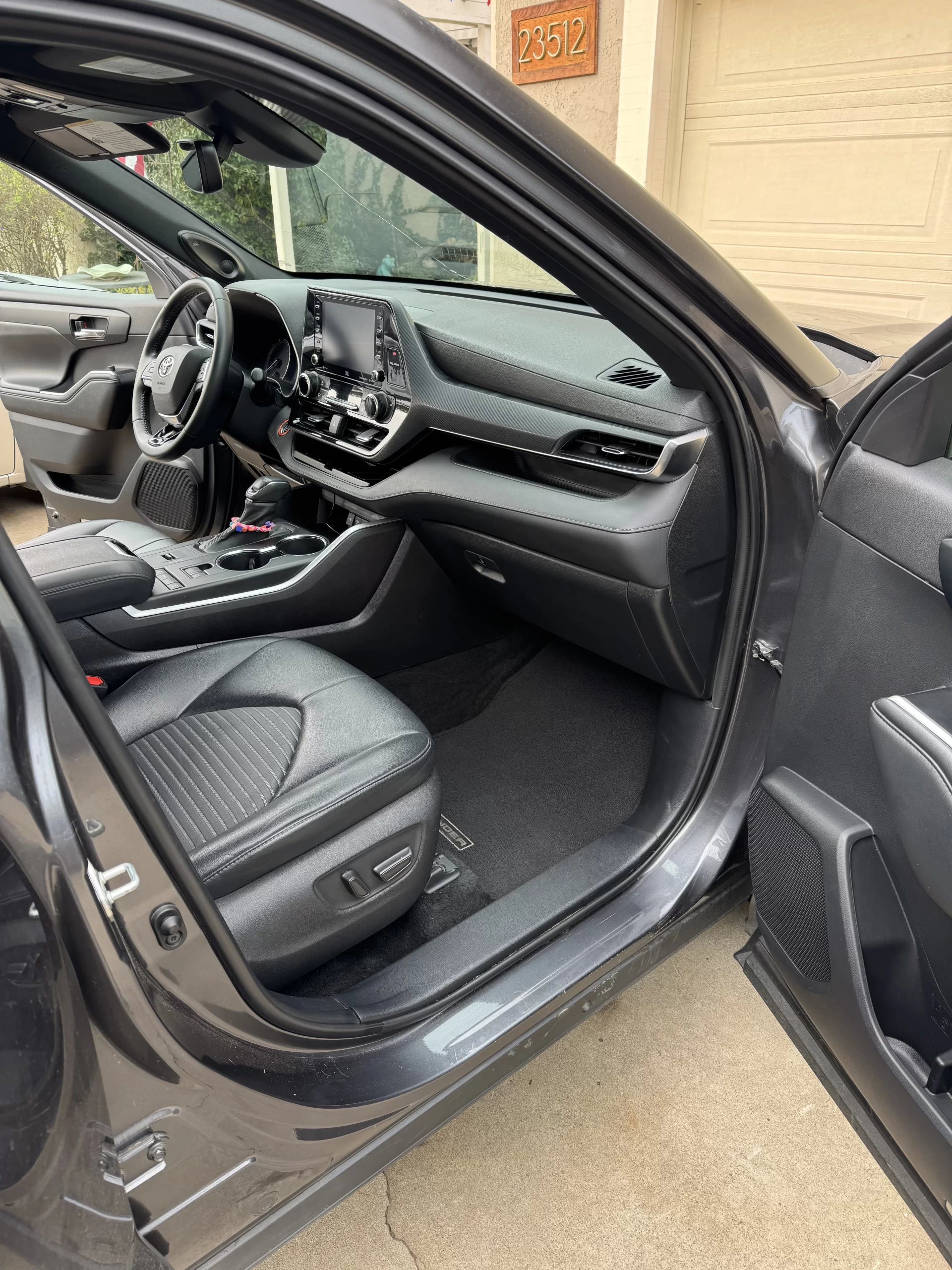 The photo shows the front passenger side of a modern gray car with the door open, revealing clean black leather seats, a touchscreen dashboard, and a tidy interior. The house number 23512 is visible outside. - DNC Auto Detailing in Ramona, California